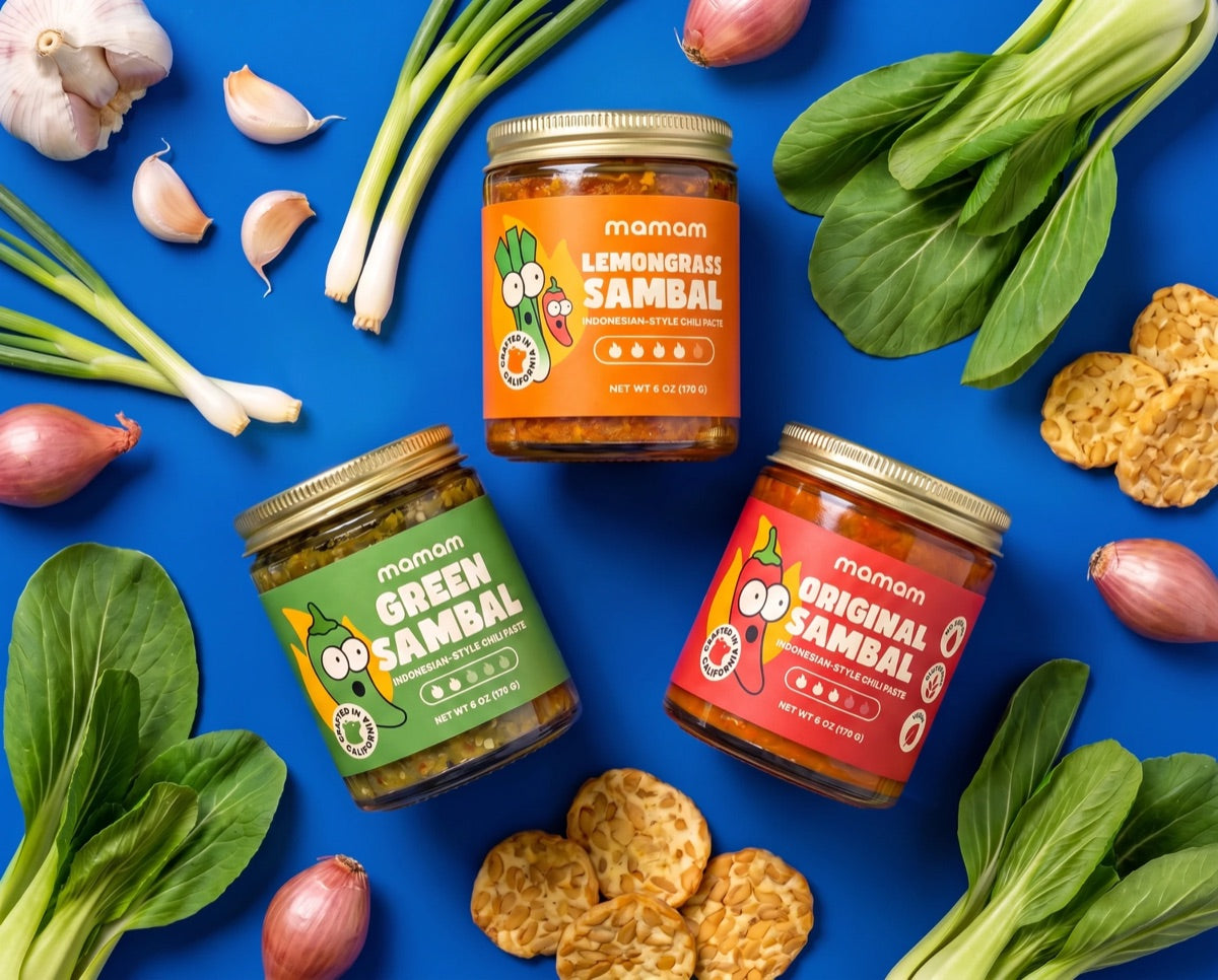 The three new MAMAM sambal jars - Lemongrass, Green and Original - on a blue table with garlic, shallots, spring onions, lemongrass, bok choy and tempeh
